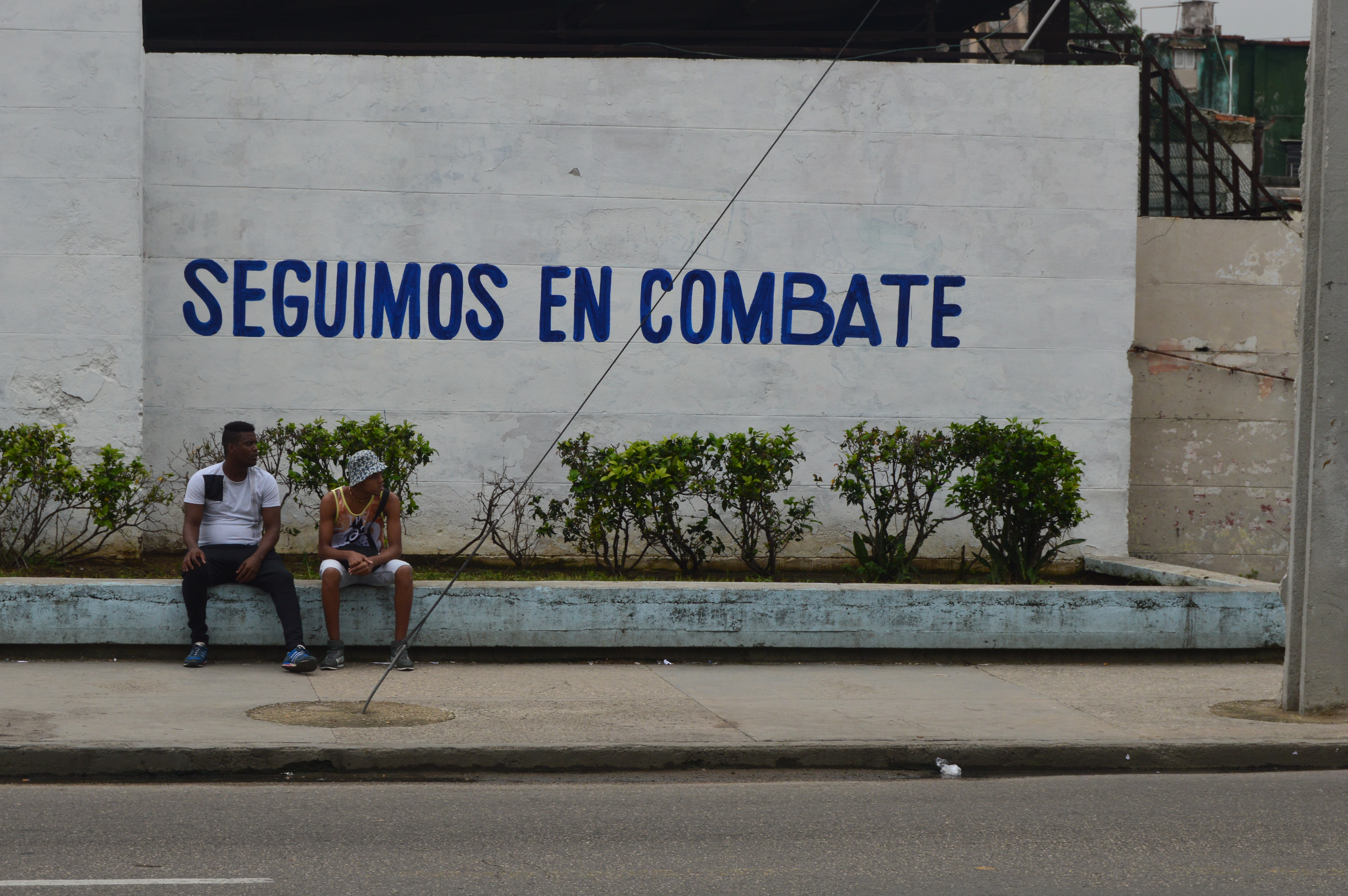 Dos personas sentadas frente a cartel revolucionario en La Habana: Seguimos en combate